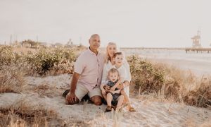 Family group sitting together on coastal dunes during golden hour with pier in background.