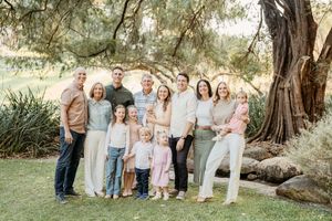 Large family group portrait smiling outdoors under a large tree in a lush green garden setting.