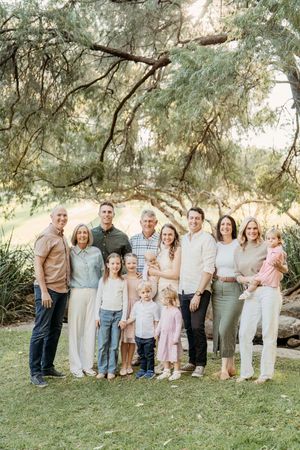 Large multi-generational family portrait outdoors under green trees, dressed in neutral tones, smiling together.