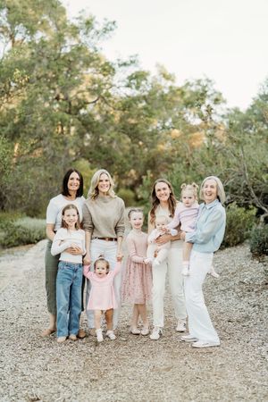 Group of women and young girls posing together on a gravel path surrounded by lush green trees outdoors.