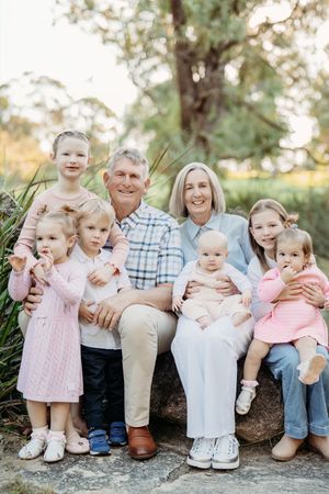 Grandparents sitting outdoors with five young grandchildren, smiling together in a lush green park setting.