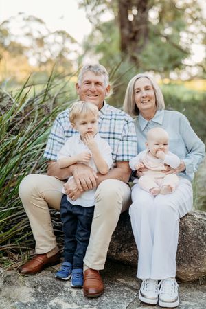 Grandparents sitting on a rock outdoors holding two young grandchildren, smiling in a lush green garden setting.