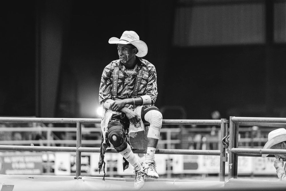 A black and white photo of a cowboy sitting on metal railings at a rodeo event.