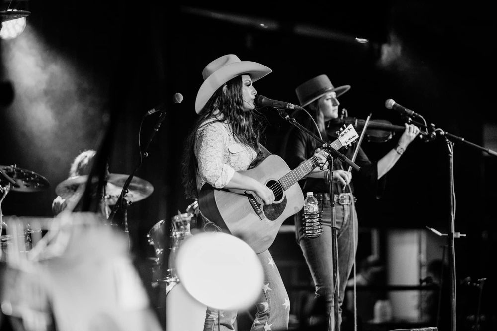 Musicians perform together on stage in black and white with acoustic guitars and stage lighting.