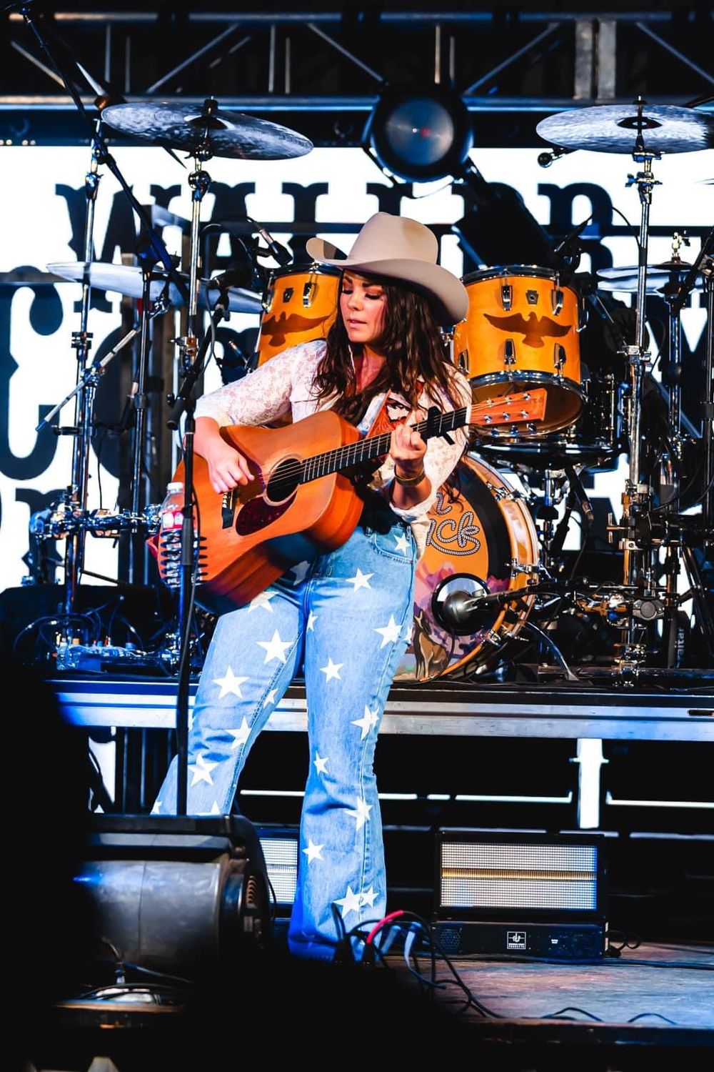 A country musician performs with an acoustic guitar on stage with orange drum kit in the background.