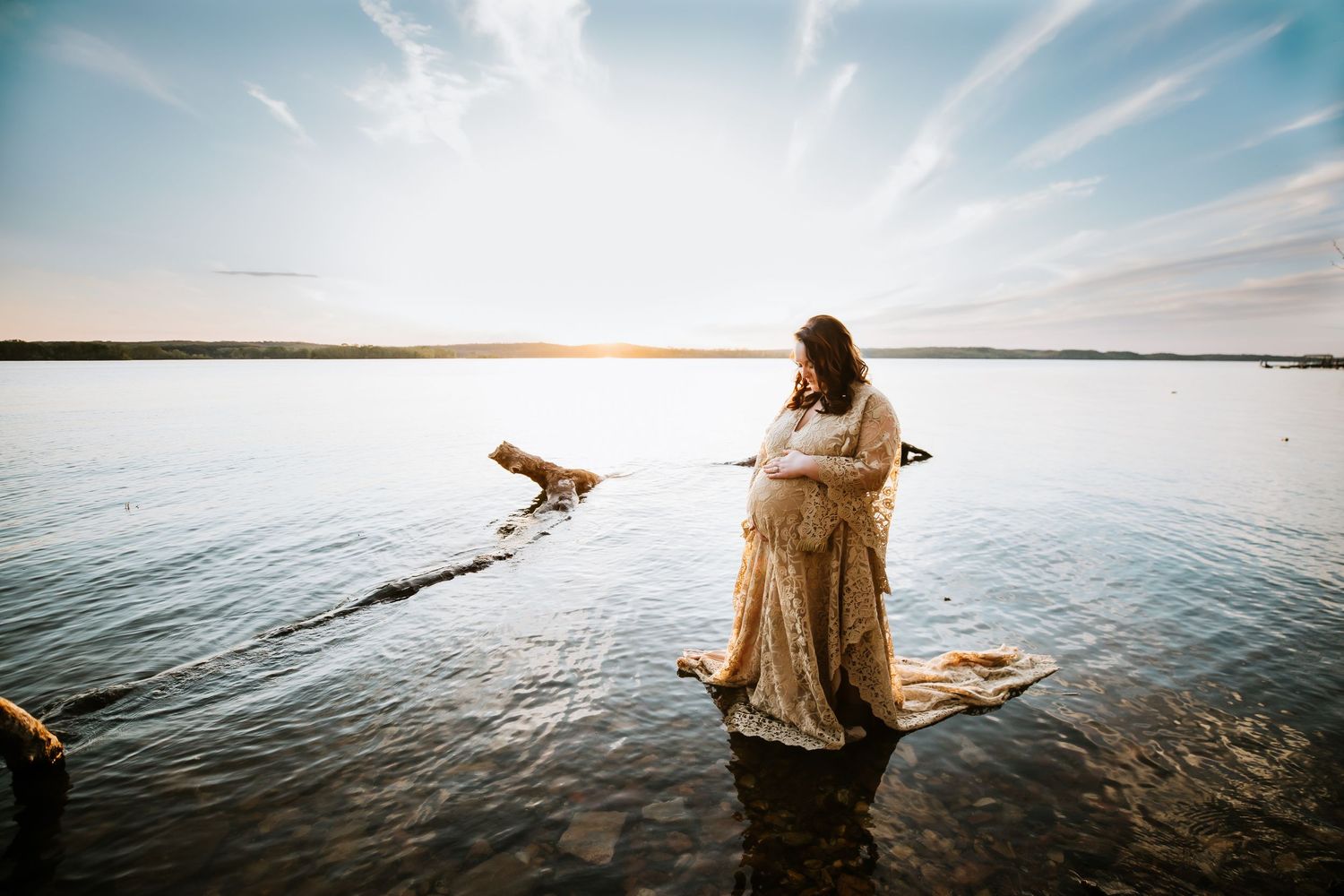 Pregnant woman stands in lake wearing cream colored gown and holding belly. Sky is blue and cloud filled at sunset in Waverly, TN