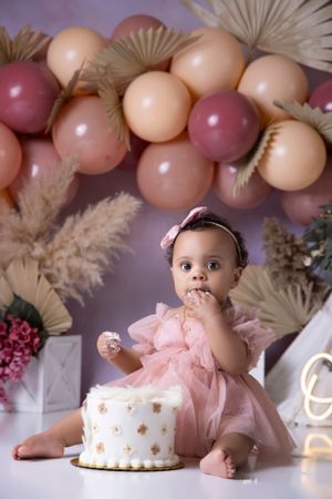 Baby in pink dress having a cake smash photo session with balloons and decorative backdrop.