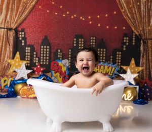 Baby in white bathtub against cityscape backdrop with stars and string lights for first birthday photoshoot.