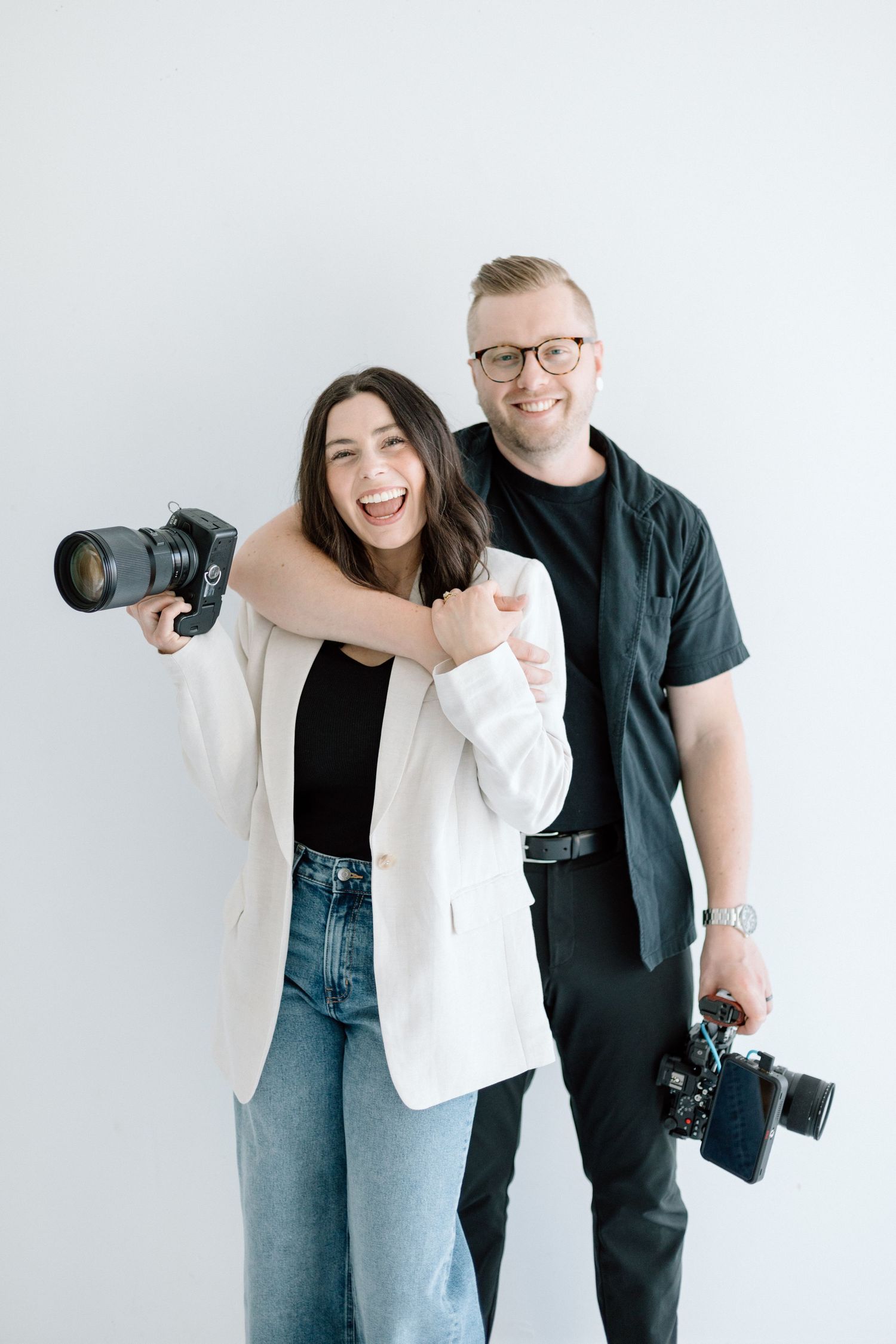 Two photographers with professional cameras pose against white backdrop.