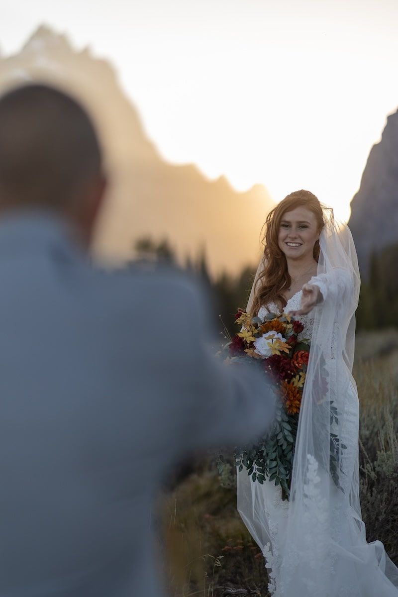 A bride in a flowing white dress and veil holds a vibrant autumn bouquet while standing in a sunlit field at sunset.