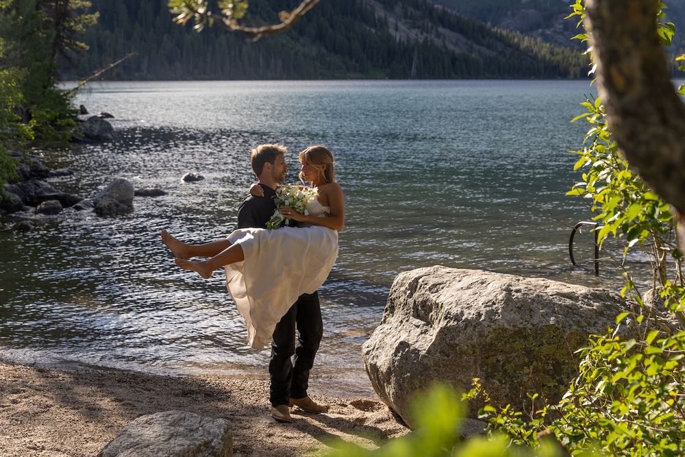 Groom carries bride in white gown by a serene mountain lake surrounded by trees and boulders on their wedding day.
