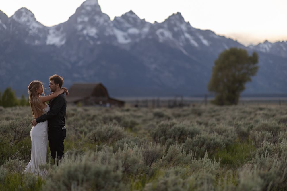 Couple embraces in a field with Grand Teton mountains and historic barn at sunset in Jackson Hole, Wyoming.
