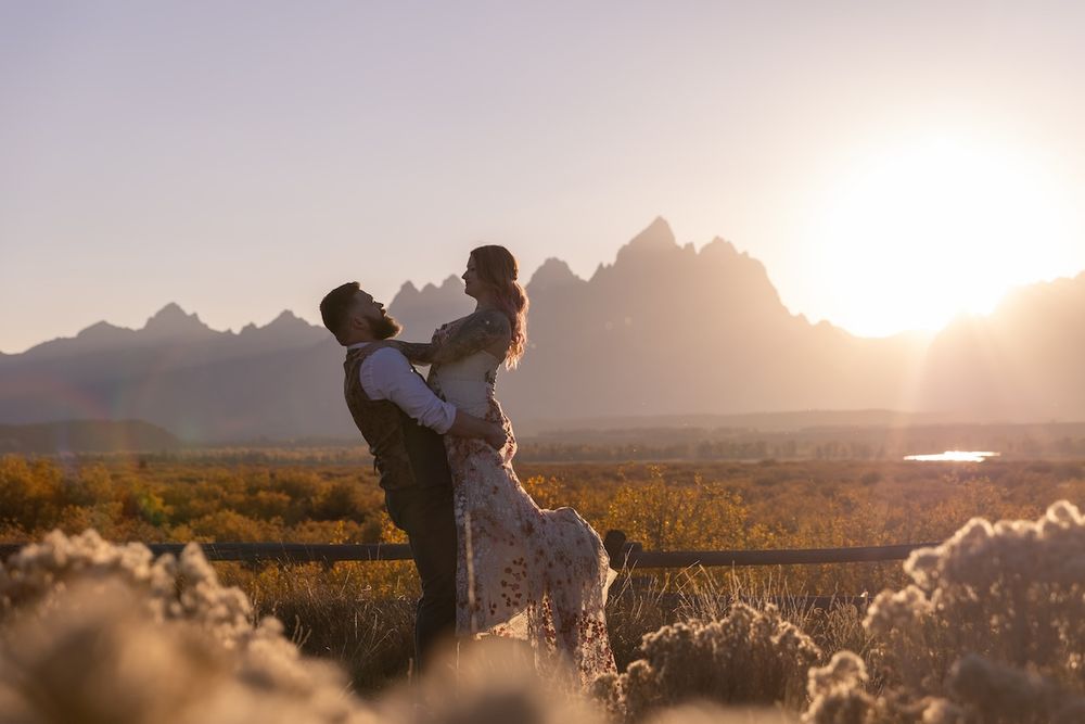 Silhouetted figure stands on mountain peak against dramatic sunset sky with mountain ranges in background.