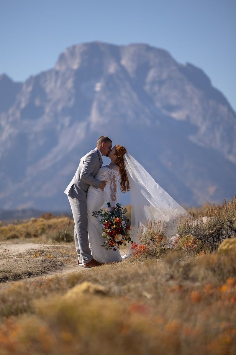 A couple in wedding attire shares an intimate moment against a backdrop of rugged mountains and wildflowers at sunset.