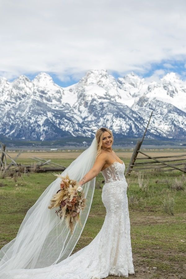 A bride in a lace wedding dress poses in front of the majestic snow-capped Grand Teton mountains on her wedding day.