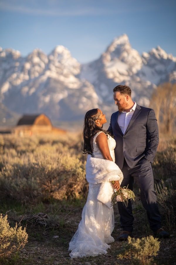 Romantic wedding couple sharing intimate moments against majestic Grand Teton mountains and rustic barn backdrop.