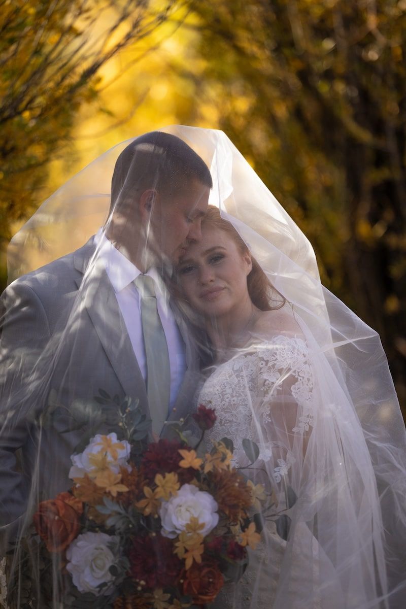 A romantic wedding couple shares an intimate moment under a veil against a backdrop of golden autumn foliage.