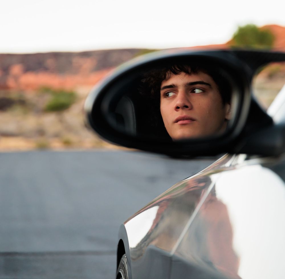 Young man's reflection in a car side mirror with scenic desert landscape in the background.