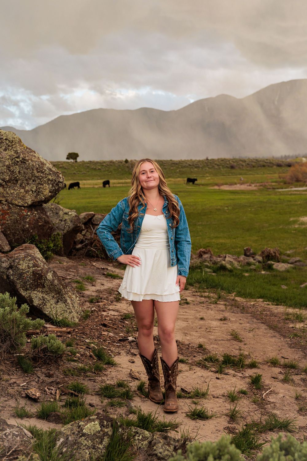 Young woman in white dress, blue jacket, and cowboy boots poses on a ranch path with mountains and cattle in background.