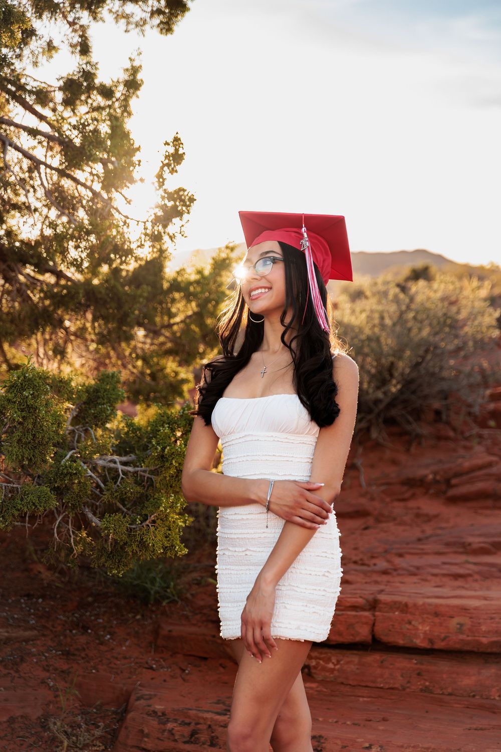 Young woman in white dress and red graduation cap smiling outdoors at sunset in red rock desert landscape.