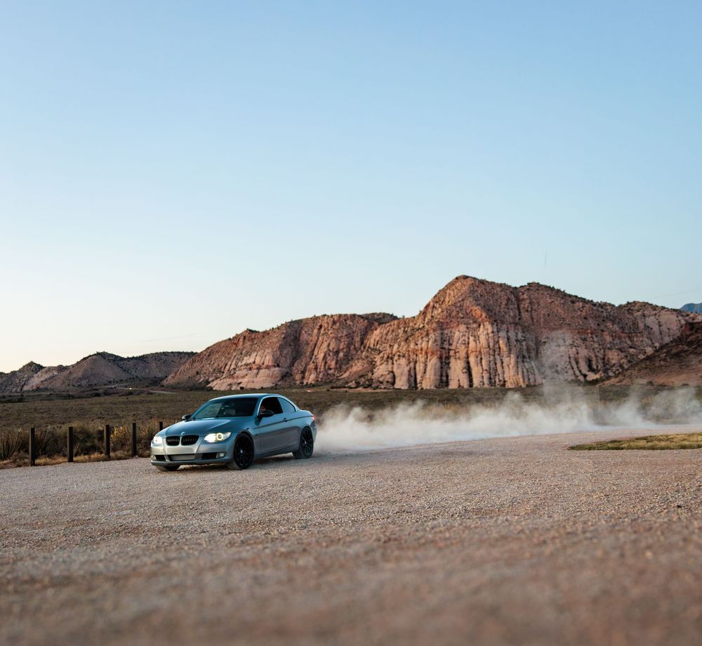 A blue BMW sedan kicks up dust on a gravel road with dramatic red rock cliffs in the desert background.