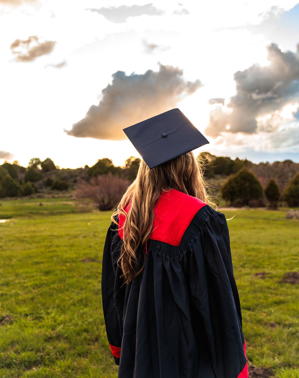 A graduate in black cap and gown with red trim stands in a green field gazing at a dramatic sunset sky.