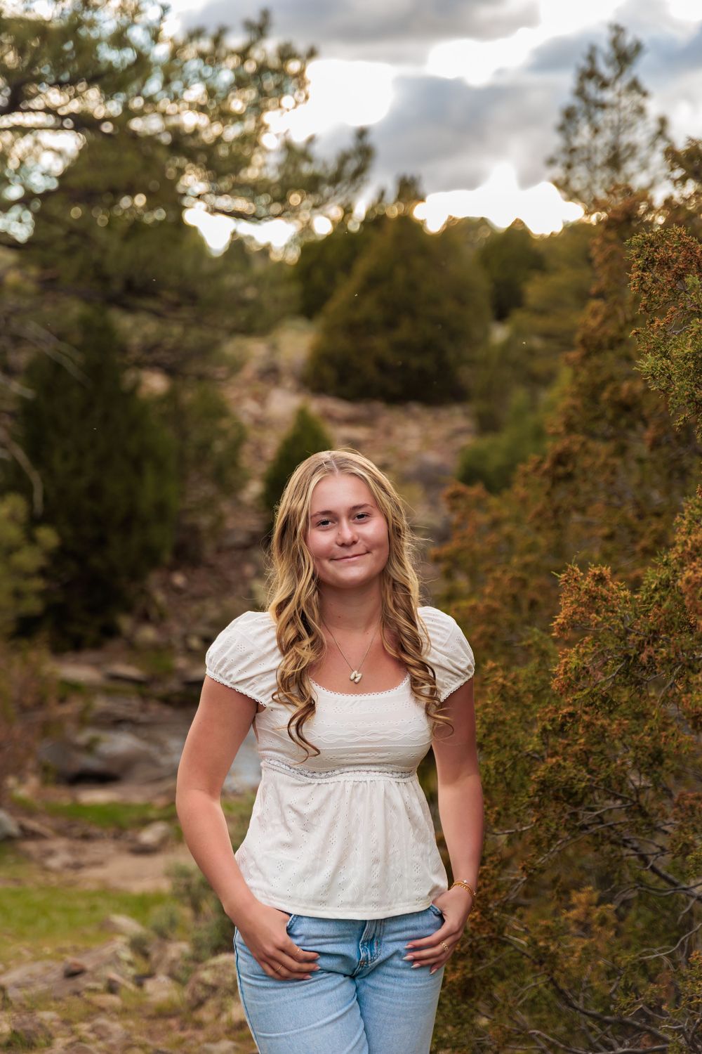 Smiling teenage girl with curly blonde hair in white top and jeans poses outdoors among pine trees at sunset.