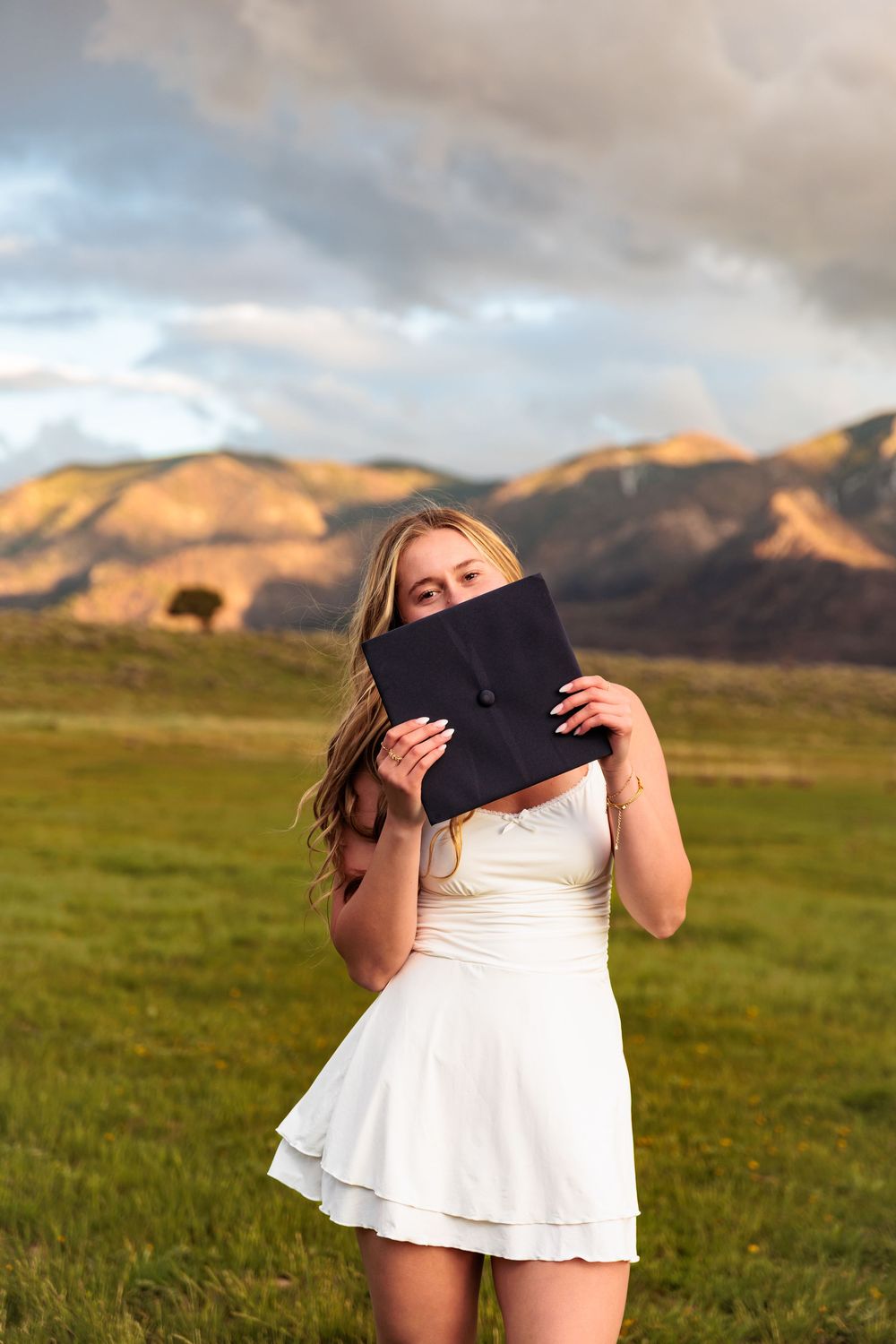 Young woman in white dress holding graduation cap over face in green field with mountain backdrop at golden hour.