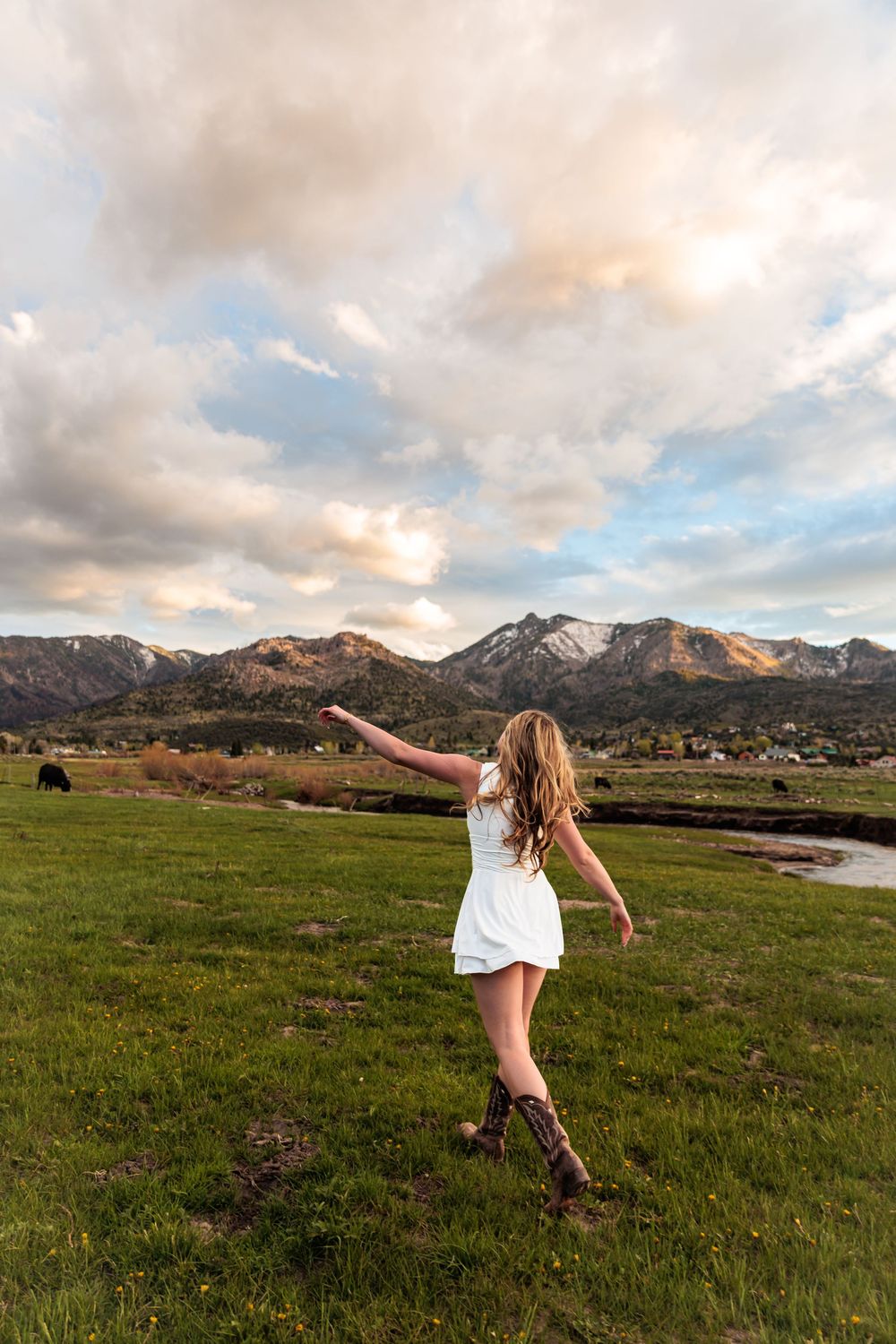 Young woman in white dress and cowboy boots twirling in a green meadow with mountain backdrop at sunset.