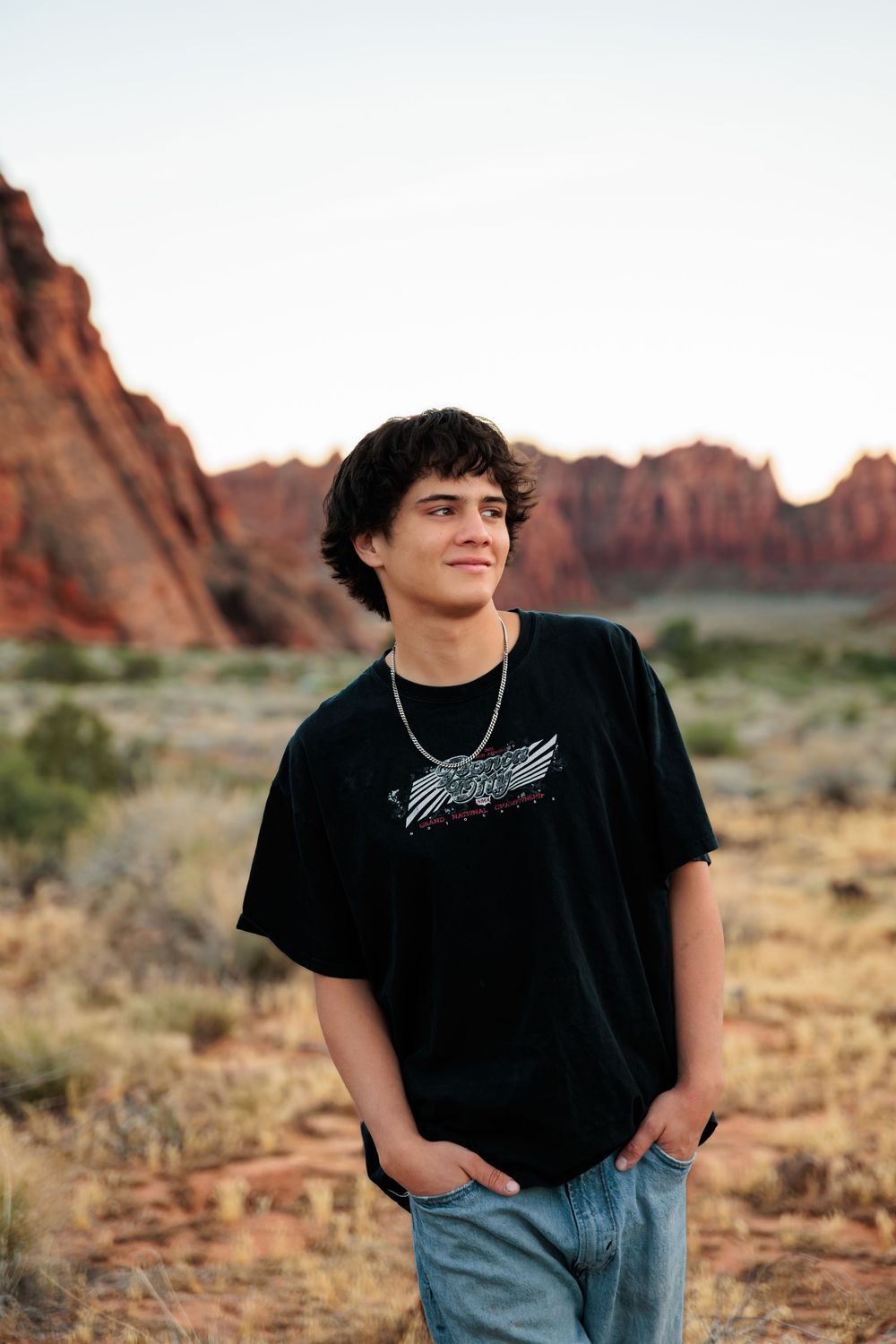 Teen boy in black graphic tee and jeans poses confidently in front of red rock desert landscape at sunset.