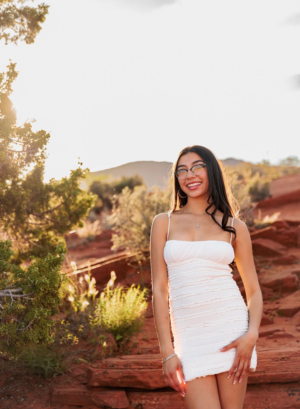 Young woman in white mini dress and glasses poses against red rock landscape at golden hour.
