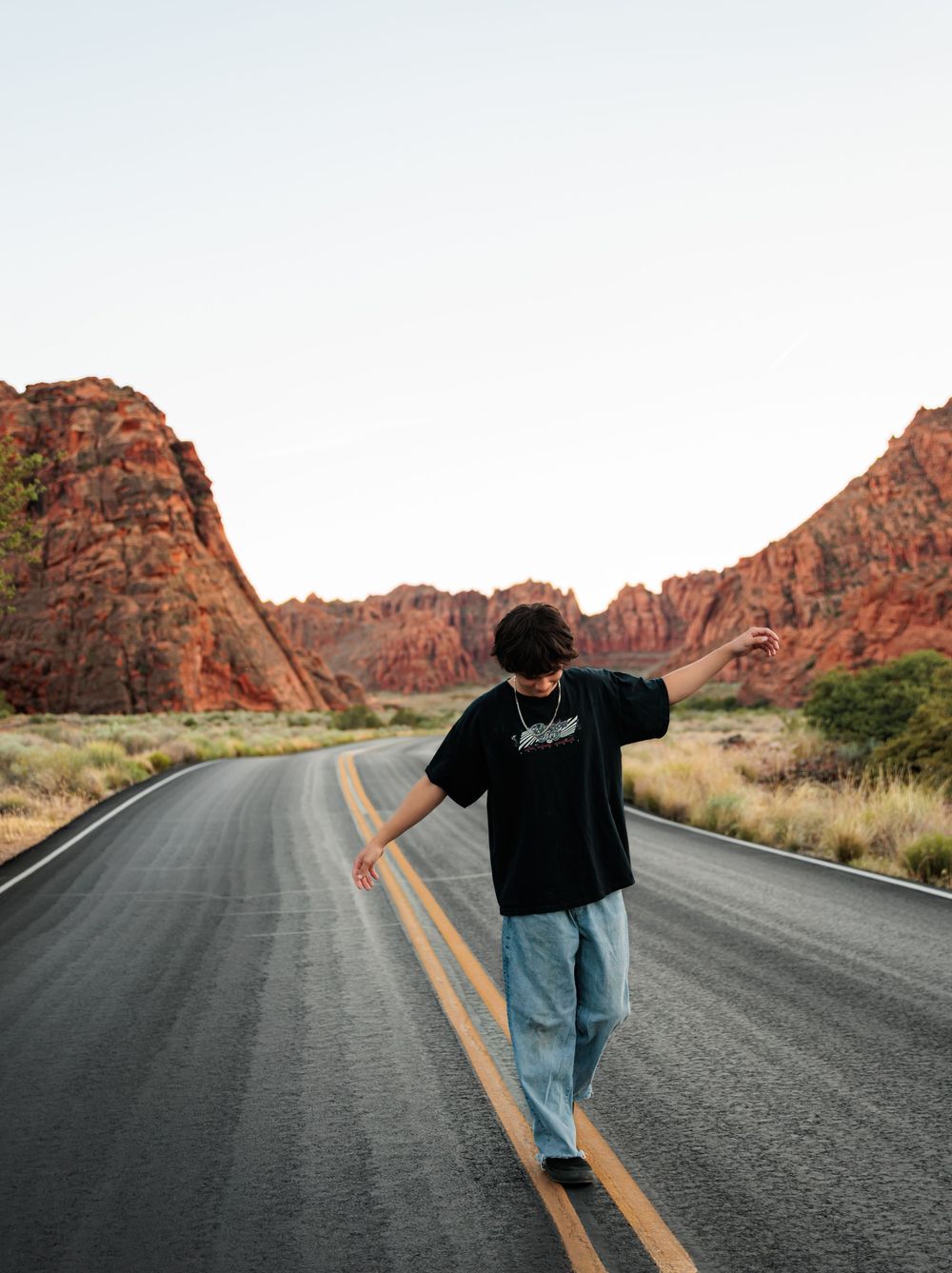 A young man in a black tee and jeans balances on a road centerline amid red rock canyon scenery.