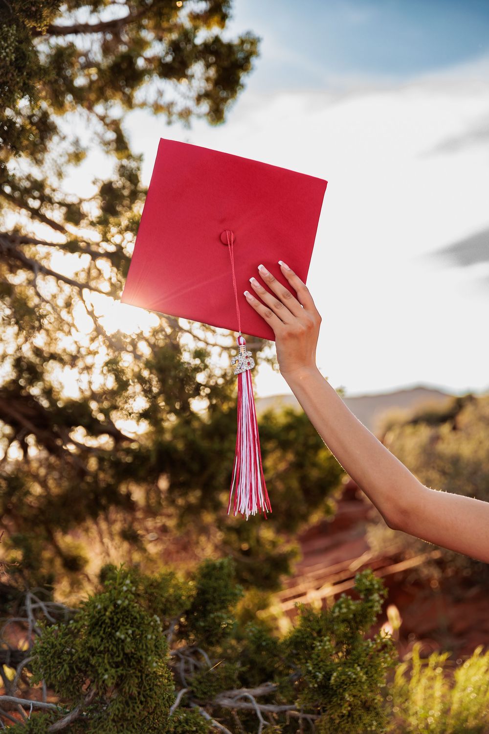A hand holds a red graduation cap with pink tassel against a sunny outdoor backdrop with trees and red rock landscape.