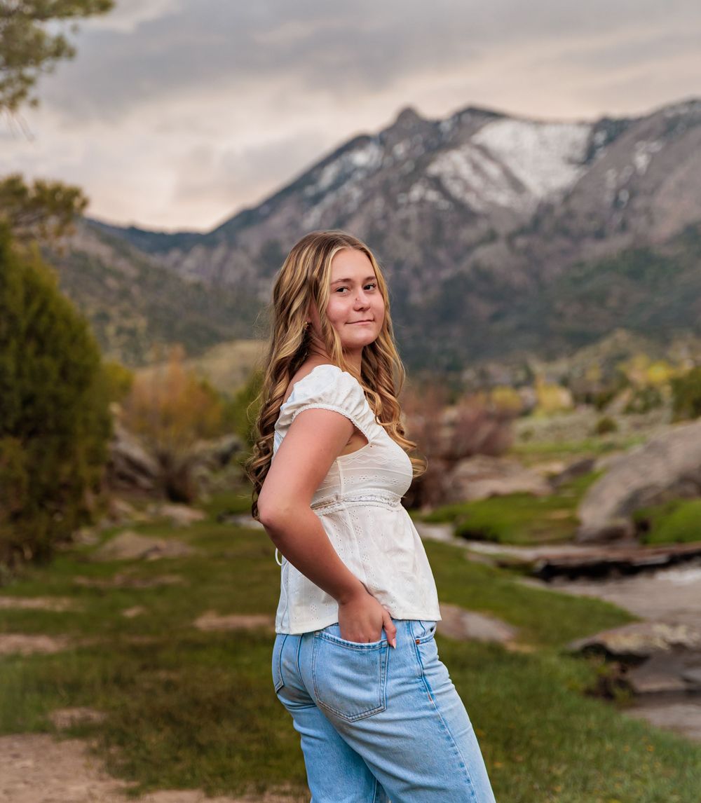 Young woman smiling in white top and jeans with Rocky Mountain backdrop and green landscape.
