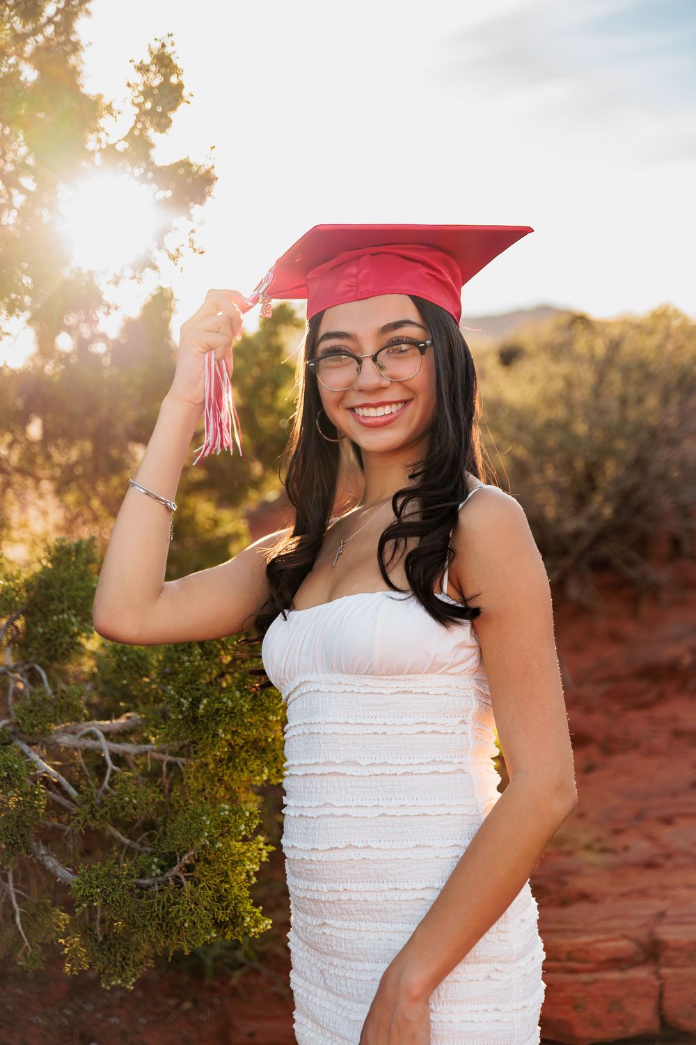 Smiling graduate in white dress and red cap holds tassel outdoors in desert landscape with golden sunlight.