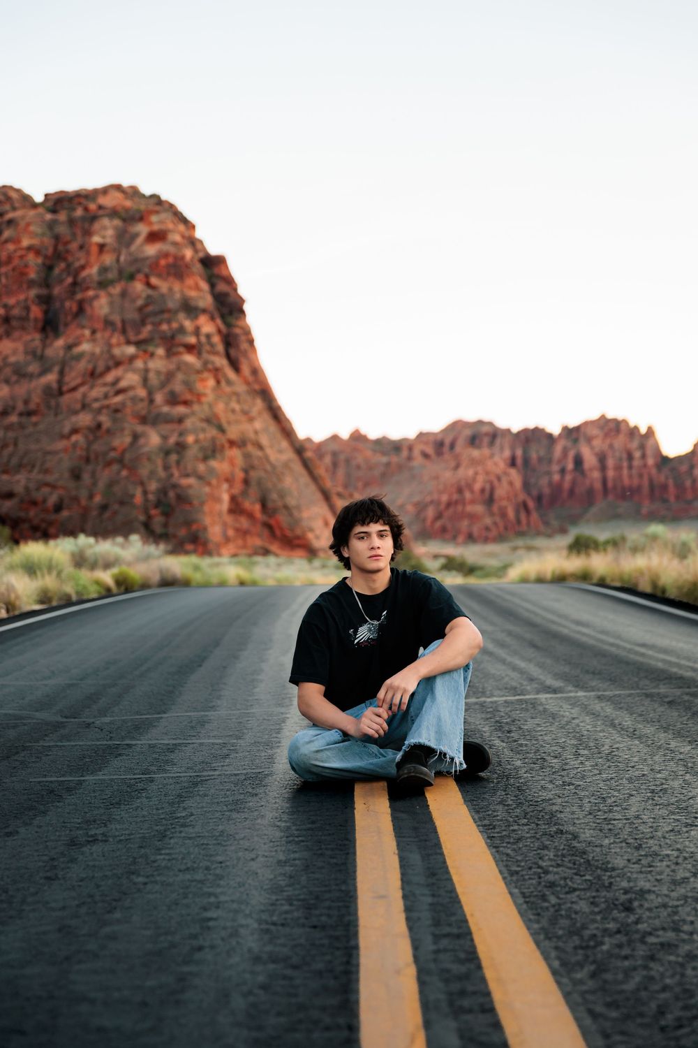 Young man sitting cross-legged on a desert highway with red rock formations in the background.