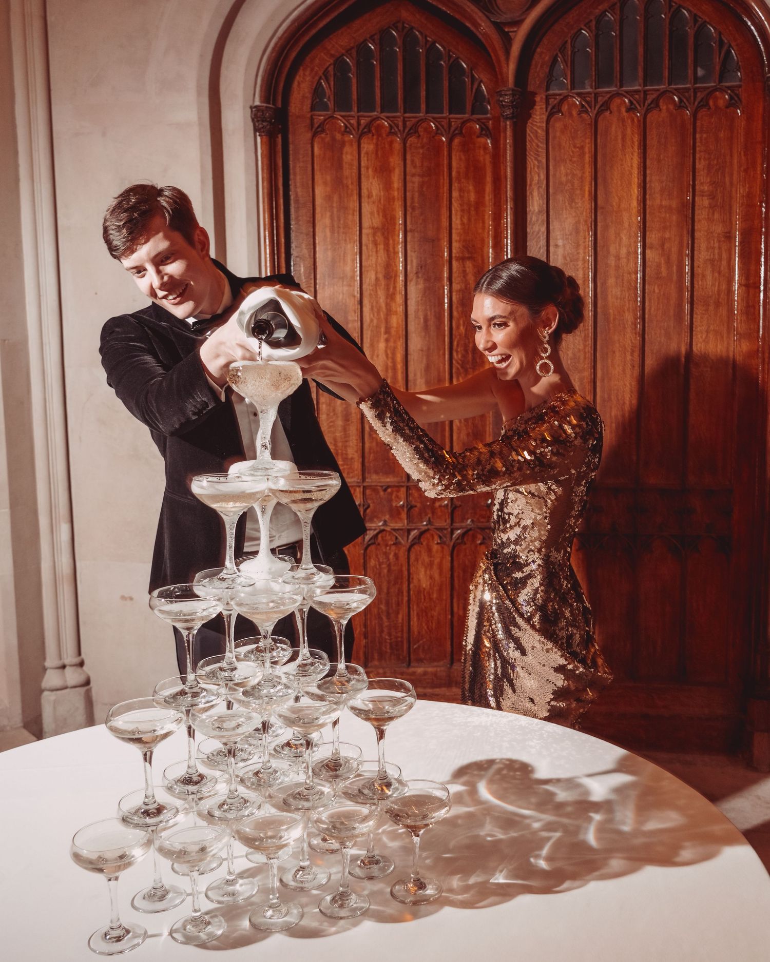 Modern wedding couple in formal attire pouring champagne into a tower of stacked crystal glasses at Ashridge House.