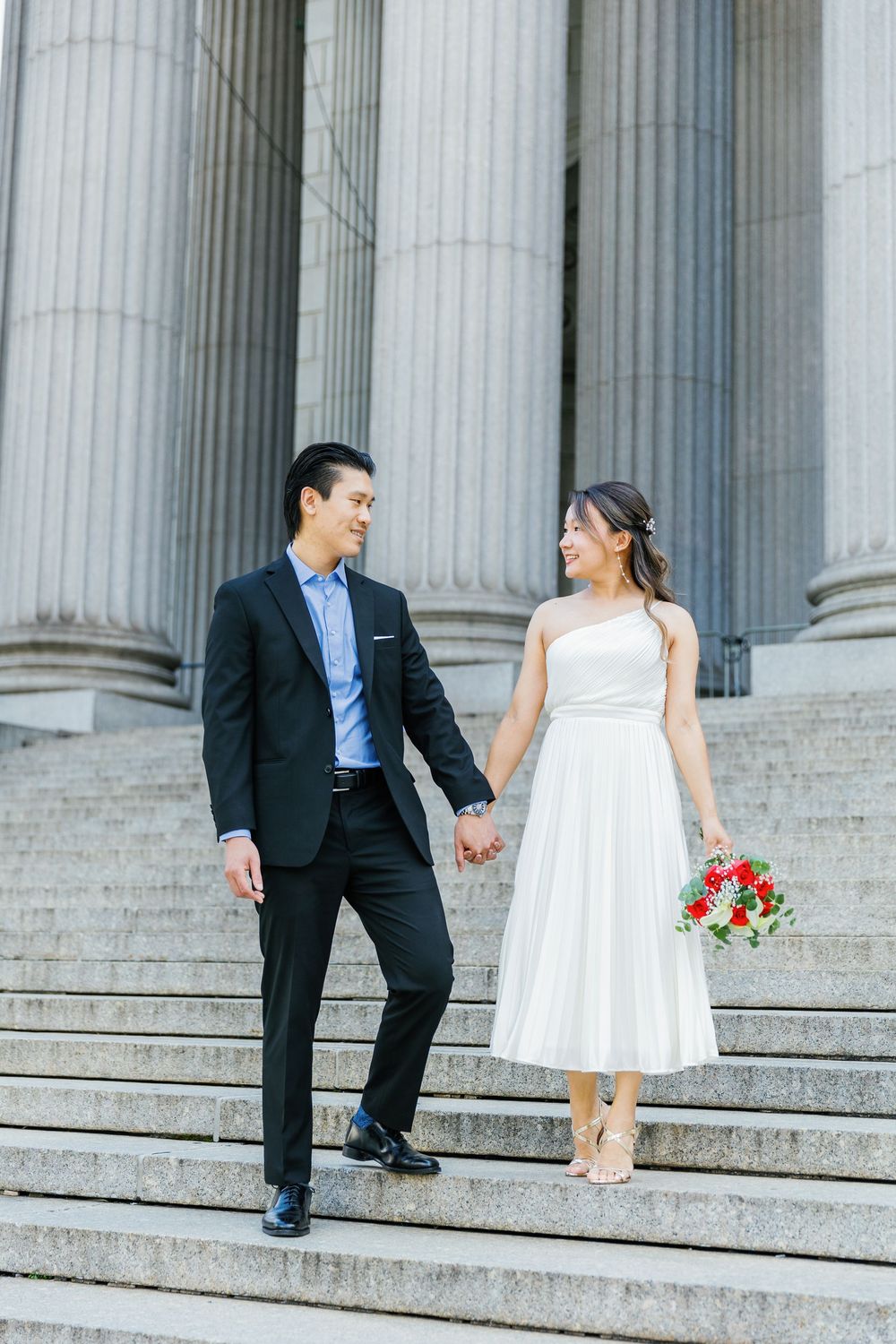 Wedding couple in white dress and black suit playfully run up courthouse steps with red bouquet and Roman columns behind.