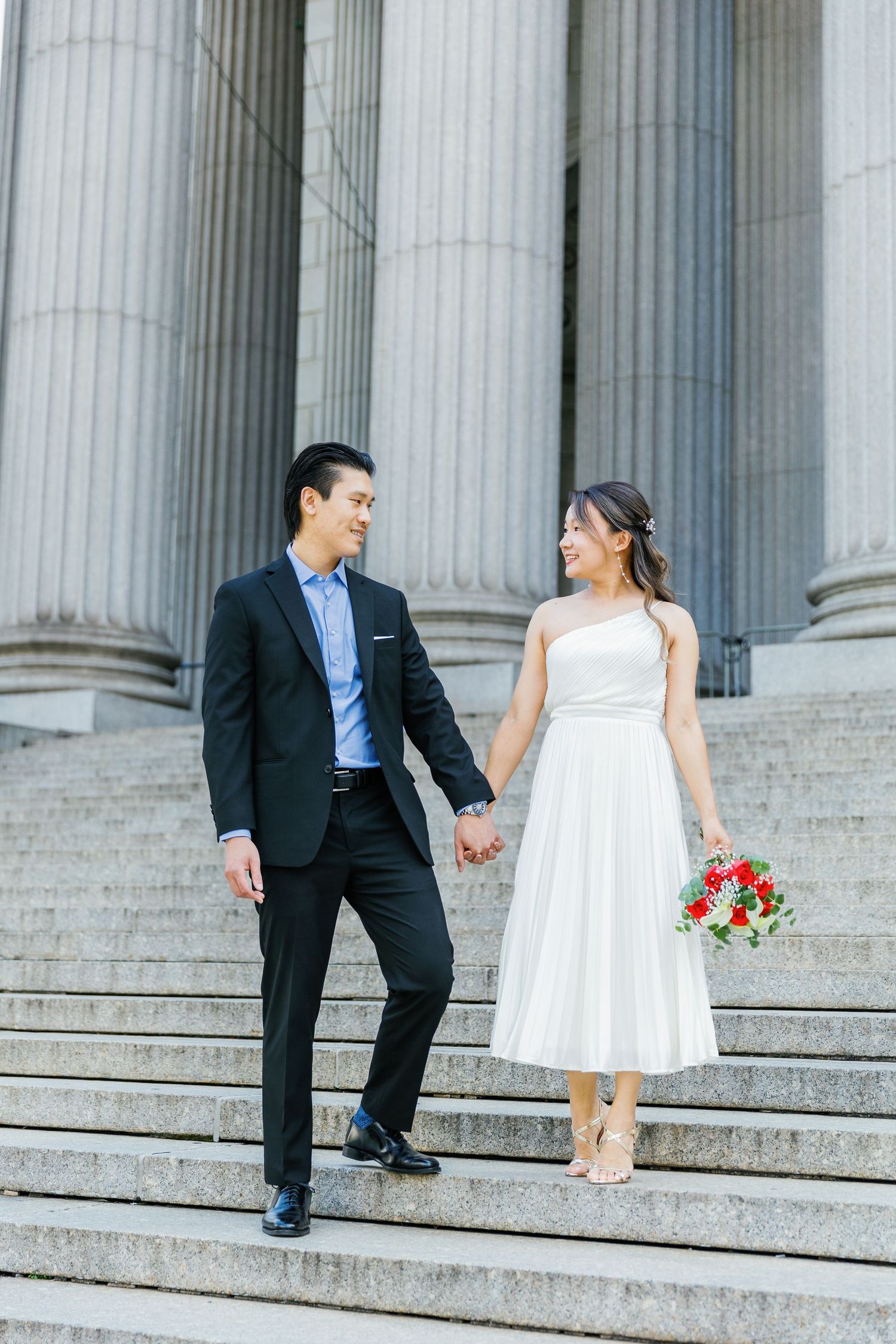 Wedding couple in white dress and black suit playfully run up courthouse steps with red bouquet and Roman columns behind.