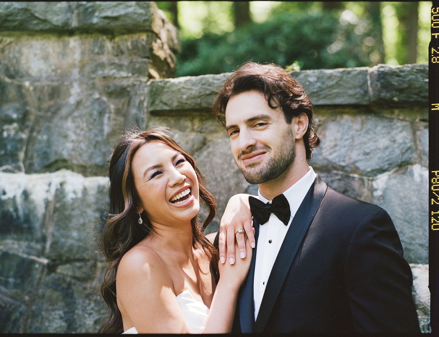 A joyful couple laughs together against a stone wall backdrop.
