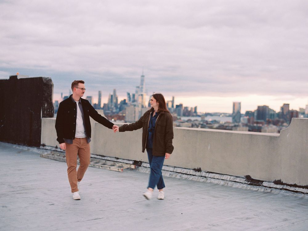 Couple holding hands on rooftop with NYC skyline in background.