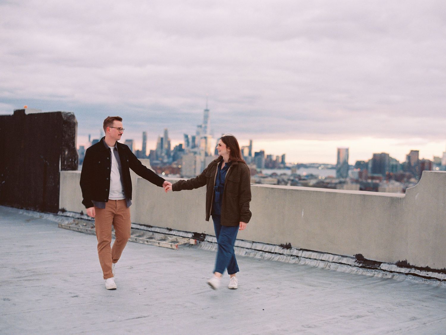 Couple holding hands on rooftop with NYC skyline in background.