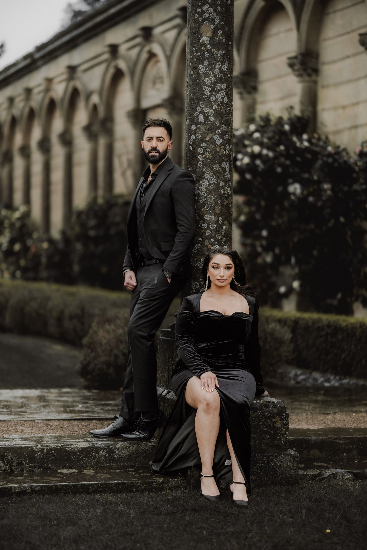 Elegant couple in black formal attire pose dramatically against gothic architectural backdrop.