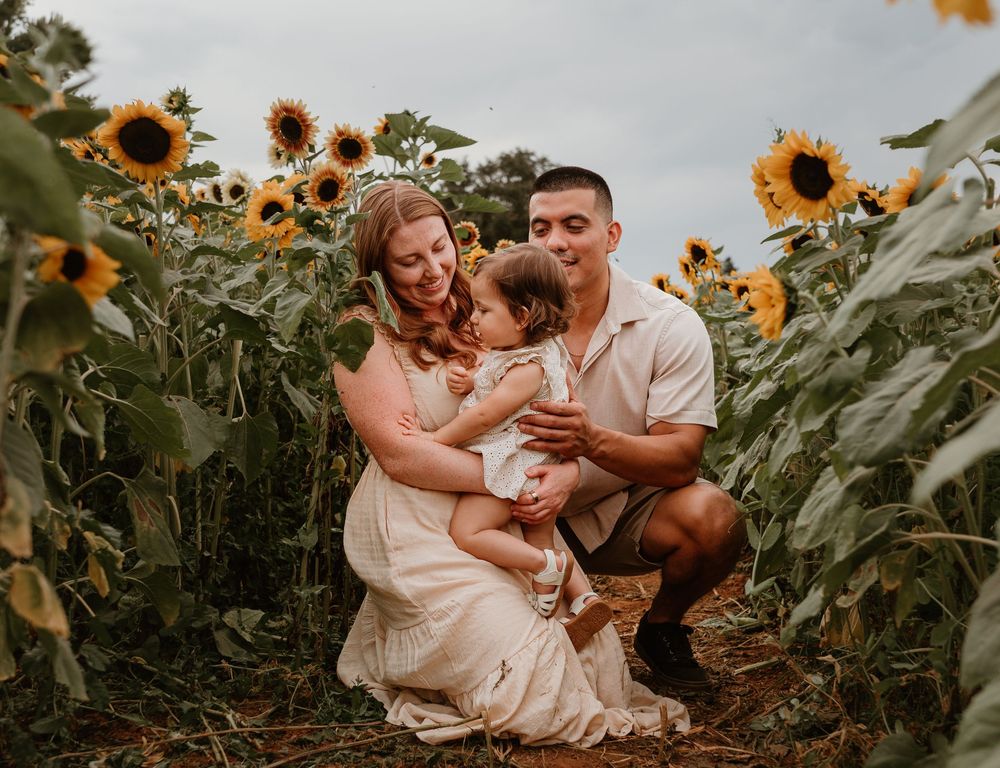 Family of three embraces and shares joyful moments while sitting among blooming sunflowers.
