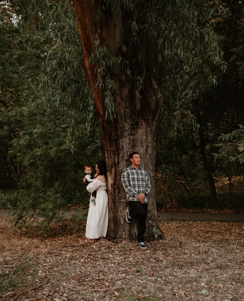 A couple stands back-to-back against a large tree trunk in a moody forest setting.