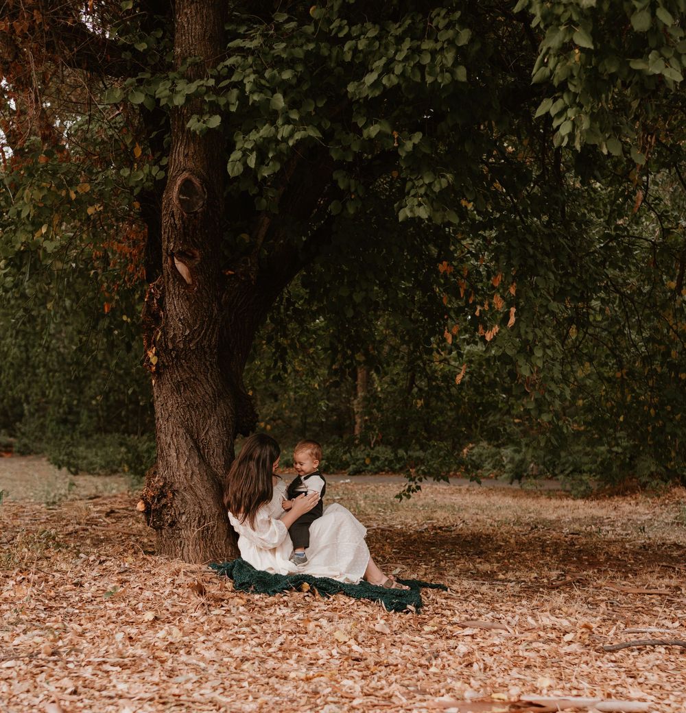 A couple sits together on fallen autumn leaves beneath a large tree in a wooded setting.