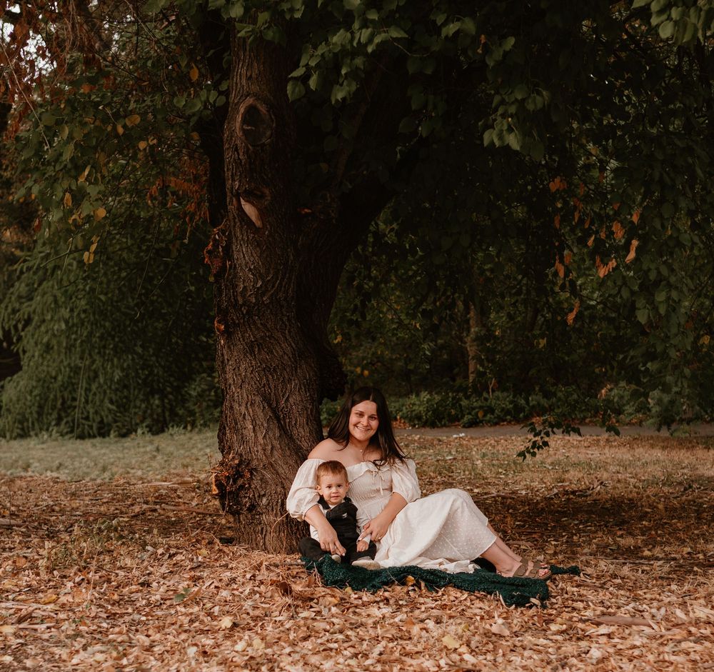 A mother and child sit together under a large tree on fallen autumn leaves in a peaceful outdoor setting.