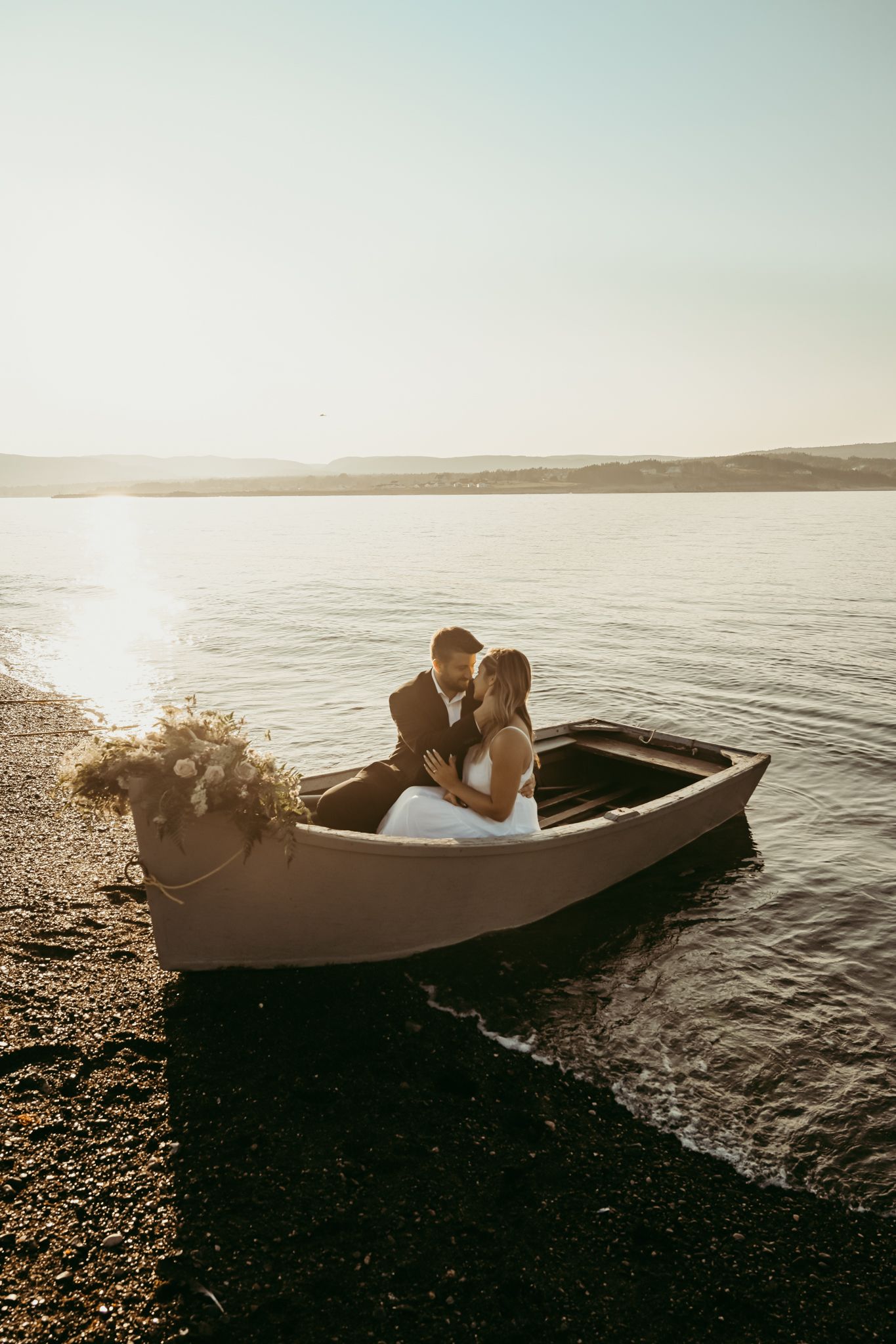 Romantic couple in wedding attire sitting in rowboat at sunset on calm waters.