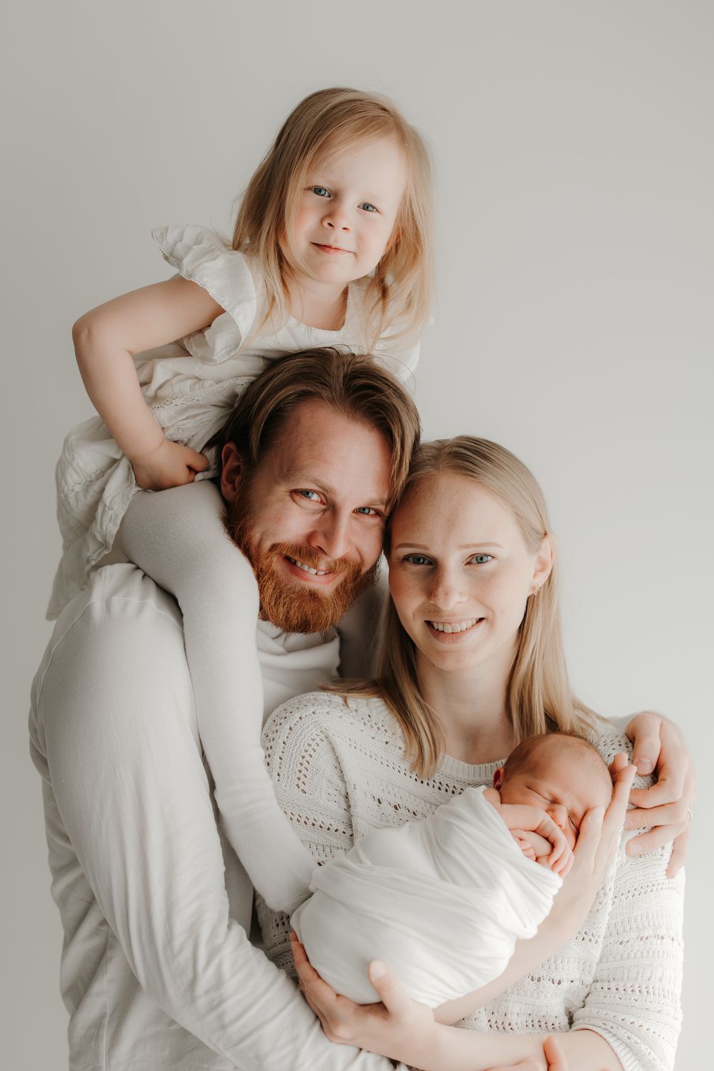 Happy family portrait with parents, toddler on dad's shoulders, and newborn baby, dressed in white.