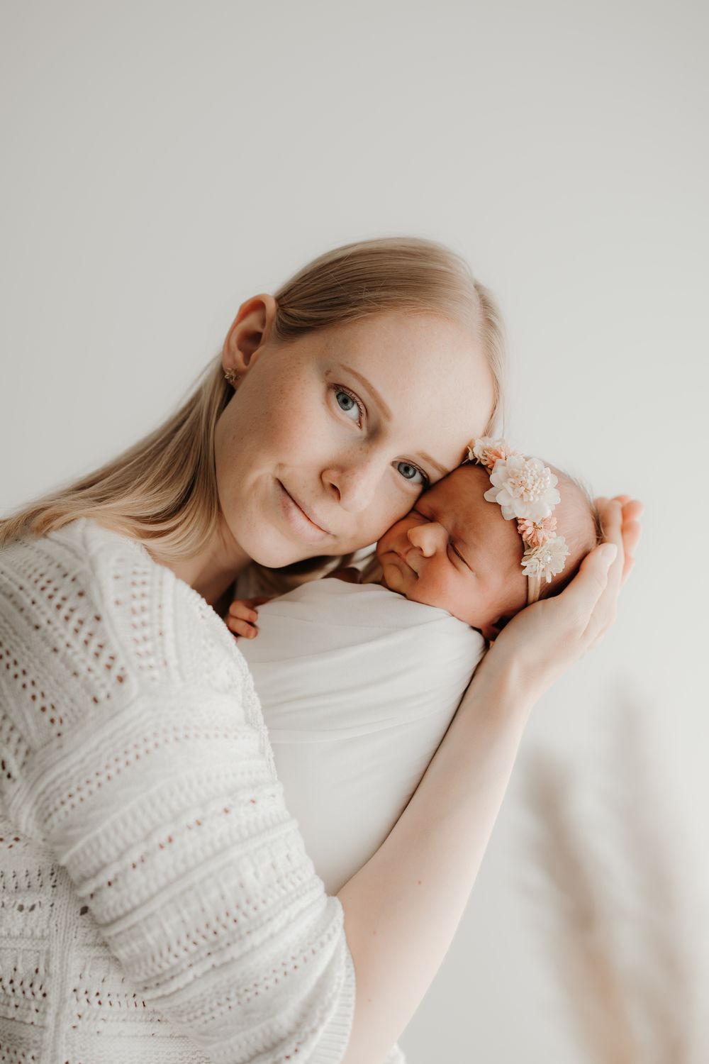 Mother in white lace top tenderly holds sleeping newborn with floral headband against her cheek.